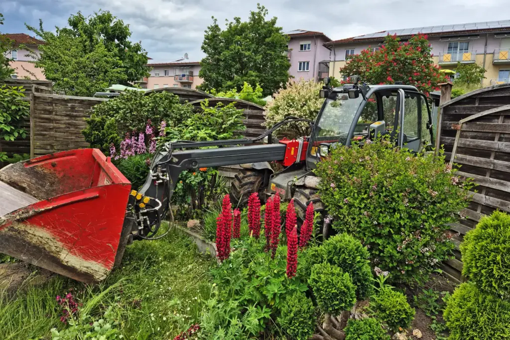 Ein Teleskoplader beim Durchqueren einer Hecke in einen Garten - im Hintergrund sieht man Häuser.