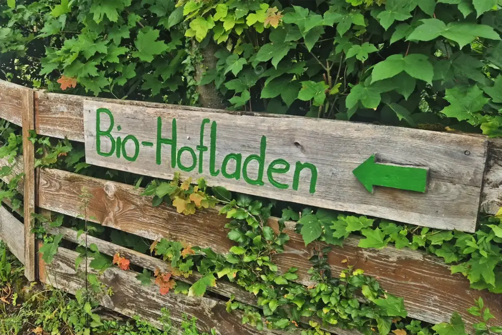 Hölzerner Wegweise mit grüner Beschriftung "Bio-Hofladen" - angebracht an einem Holzzaun, dahinter Blattwerk